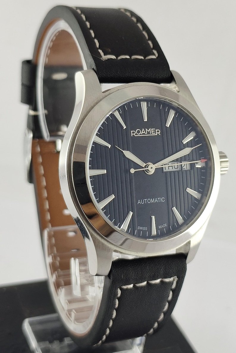 May include: A Roamer automatic wristwatch with a dark blue dial and silver hands. The watch features a black leather strap with white stitching. The watch face displays the day and date. The watch is Swiss made.