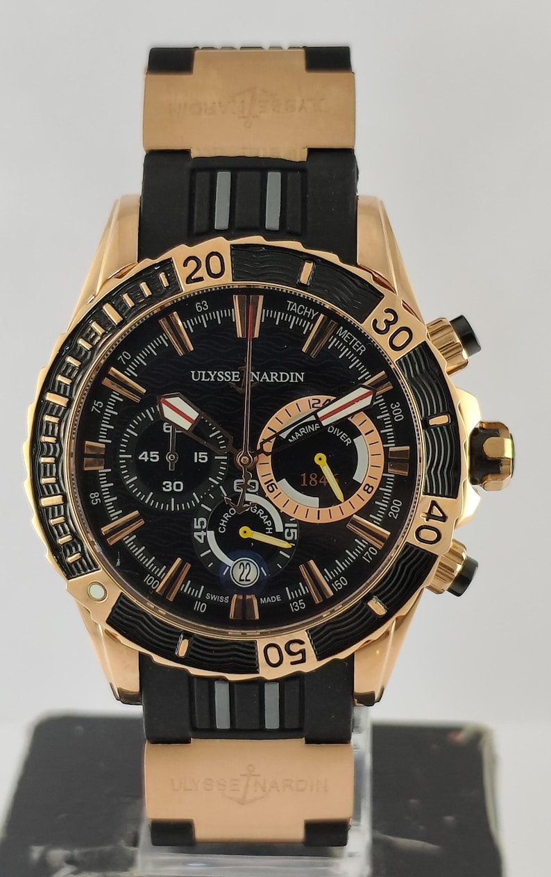 May include: A rose gold and black Ulysse Nardin Marine Diver chronograph watch with a black rubber strap. The black dial features a wave pattern, luminous hands, three sub-dials, a date window at the six o'clock position, and tachymeter markings.