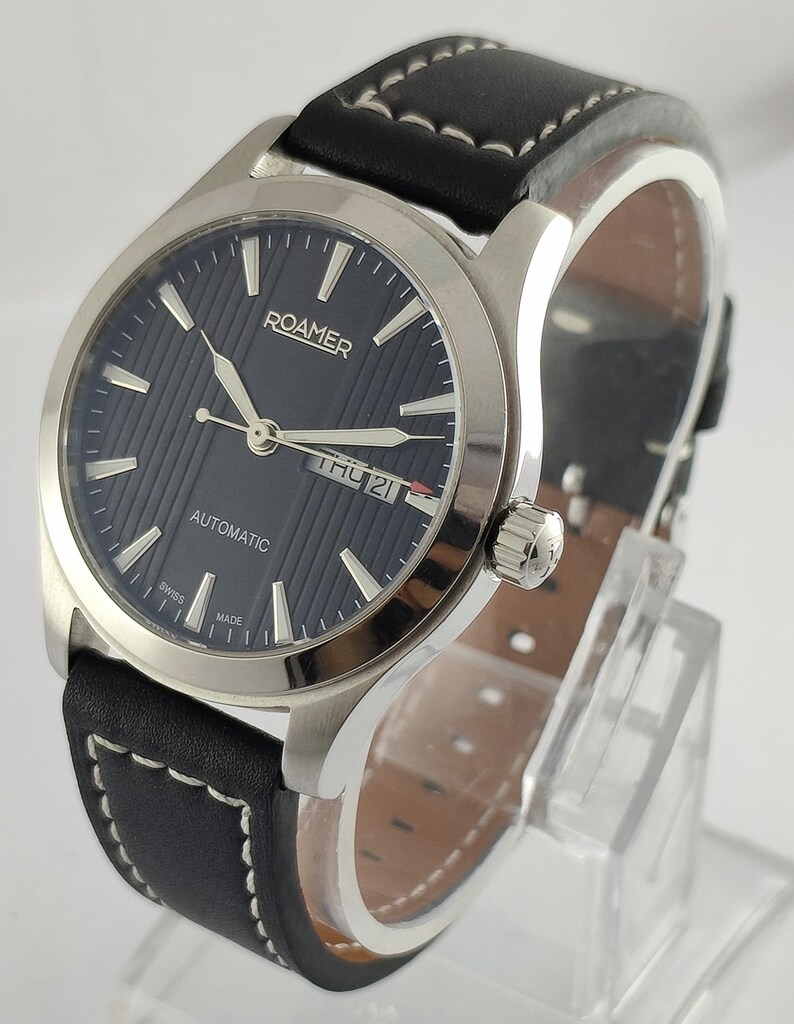 May include: A Roamer automatic wristwatch with a black leather band and silver-tone case. The watch face is dark blue with silver hands and markers. The day and date are displayed at the 3 o'clock position. The band is stitched with white thread.