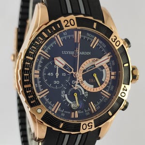 May include: A Ulysse Nardin Marine Diver chronograph watch with a rose gold case, a dark blue dial, and a black rubber strap. The watch features luminous hands, a date window at the six o'clock position, and a rotating black bezel with gold numerals.