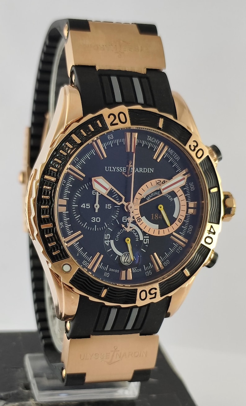 May include: A Ulysse Nardin Marine Diver chronograph watch with a rose gold case, a dark blue dial, and a black rubber strap. The watch features luminous hands, a date window at the six o'clock position, and a rotating black bezel with gold numerals.