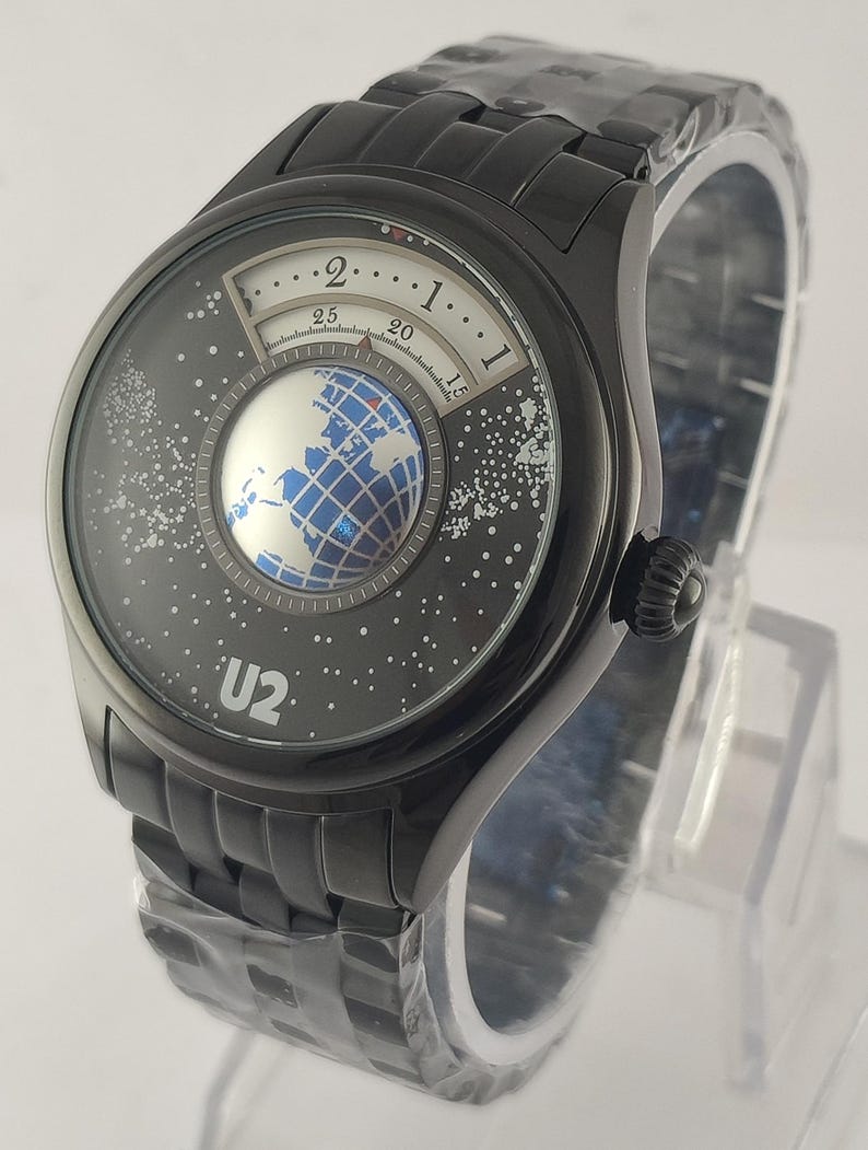 Puede incluir: Reloj de pulsera negro con un dise&ntilde;o de esfera &uacute;nico. La esfera del reloj presenta un gr&aacute;fico de globo terr&aacute;queo y un fondo estrellado, con el texto "U2". El reloj tiene una correa de metal negro y un bisel negro.