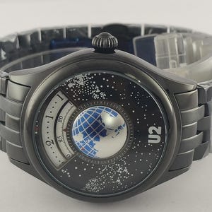 Puede incluir: Un reloj de pulsera gris oscuro con un dise&ntilde;o &uacute;nico. La esfera del reloj presenta un globo azul y plateado, un fondo estrellado y el texto "U2". El reloj tiene una correa gris oscuro a juego.