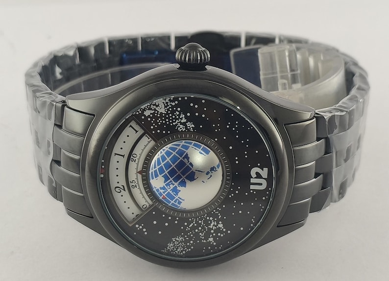 Puede incluir: Un reloj de pulsera gris oscuro con un dise&ntilde;o &uacute;nico. La esfera del reloj presenta un globo azul y plateado, un fondo estrellado y el texto "U2". El reloj tiene una correa gris oscuro a juego.
