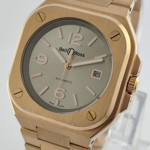 May include: A gold-toned, square-faced wristwatch with a matching band. The watch face is light gray with gold-toned numerals and hands. The brand name "Bell & Ross" is displayed above the center. The watch is labeled "Automatic" and "Swiss Made".