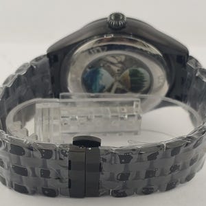 Puede incluir: Un reloj de pulsera negro con una correa de acero inoxidable. La esfera del reloj es visible a trav&eacute;s de la parte trasera transparente, revelando el mecanismo interno. La correa est&aacute; hecha de eslabones entrelazados y tiene un cierre.