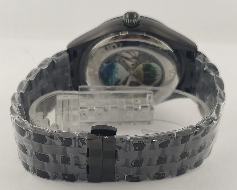 Puede incluir: Un reloj de pulsera negro con una correa de acero inoxidable. La esfera del reloj es visible a trav&eacute;s de la parte trasera transparente, revelando el mecanismo interno. La correa est&aacute; hecha de eslabones entrelazados y tiene un cierre.