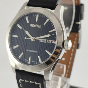 May include: A silver Roamer automatic watch with a dark blue dial and a black leather strap. The watch face displays the day and date, with white hands and markers. The strap has white stitching.