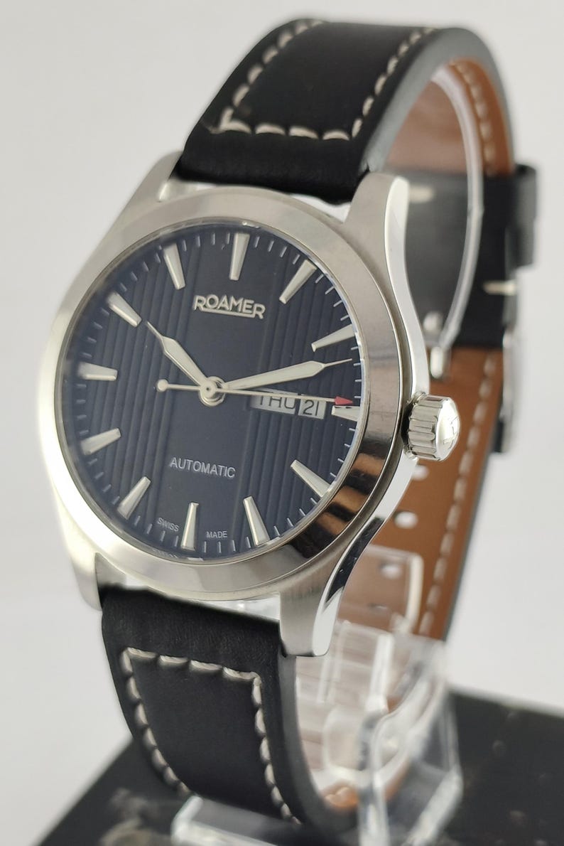 May include: A silver Roamer automatic watch with a dark blue dial and a black leather strap. The watch face displays the day and date, with white hands and markers. The strap has white stitching.