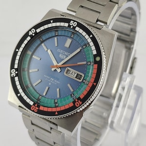 May include: A stainless steel Seiko 5 watch with a blue dial, featuring a day-date display and a rotating black bezel with white markings. The watch has a metal band and is displayed on a clear stand.