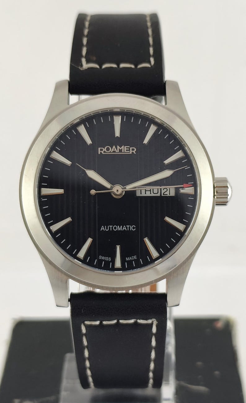 May include: A Roamer automatic wristwatch with a black dial and silver hands. The watch has a stainless steel case and a black leather strap with white stitching. The day and date are displayed at the 3 o'clock position. The watch is Swiss made.