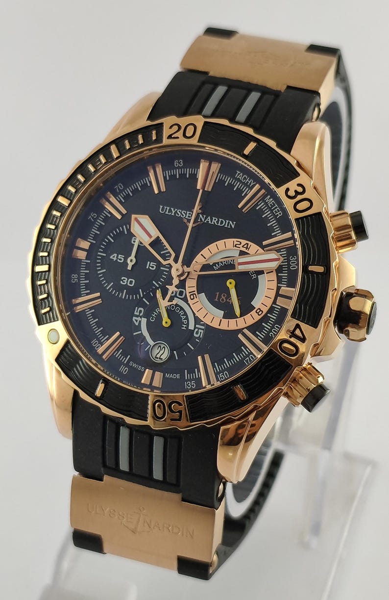 May include: A rose gold Ulysse Nardin Marine Chronograph watch with a dark blue dial, black rubber strap, and gold accents. The watch features a tachymeter scale, sub-dials, a date window at the six o'clock position, and luminous hands.