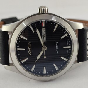 May include: A Roamer automatic wristwatch with a black dial and silver-tone hands and markers. The watch has a stainless steel case and a black leather strap with white stitching. The date is displayed at the 3 o'clock position.