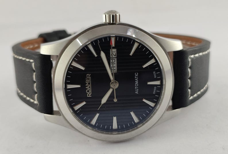 May include: A Roamer automatic wristwatch with a black dial and silver-tone hands and markers. The watch has a stainless steel case and a black leather strap with white stitching. The date is displayed at the 3 o'clock position.