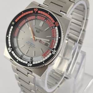 Może przedstawiać: Srebrny zegarek Seiko 5 Sports z szarą tarczą, czarnym i czerwonym pierścieniem oraz bransoletą ze stali nierdzewnej. Tarcza zegarka wyświetla dzień i datę. Zegarek ma wodoszczelność 70 metrów i 21 kamieni.