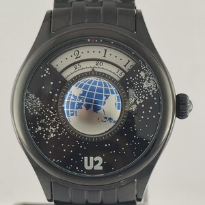 Puede incluir: Reloj de pulsera negro con un dise&ntilde;o &uacute;nico. La esfera del reloj presenta un globo terr&aacute;queo y un fondo estrellado, con el nombre de la banda "U2". El reloj tiene una correa y una caja de metal negro.