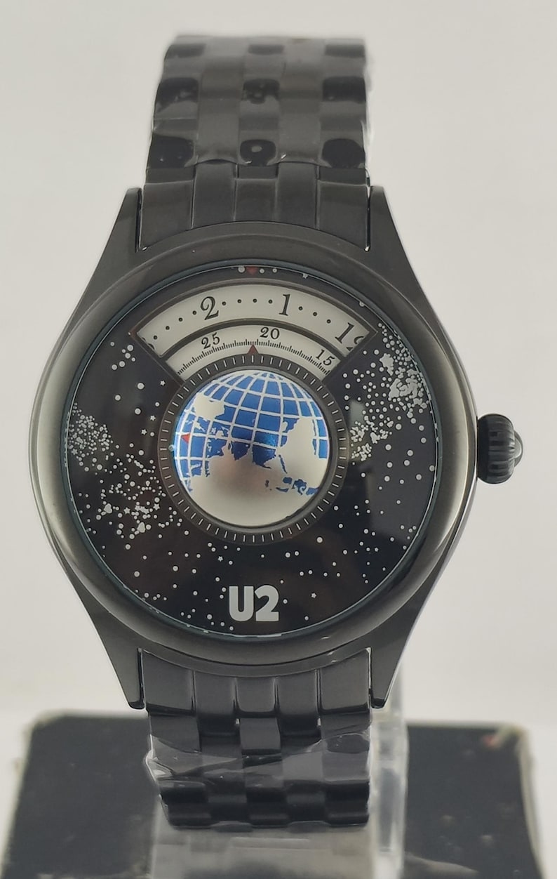 Puede incluir: Reloj de pulsera negro con un dise&ntilde;o &uacute;nico. La esfera del reloj presenta un globo terr&aacute;queo y un fondo estrellado, con el nombre de la banda "U2". El reloj tiene una correa y una caja de metal negro.