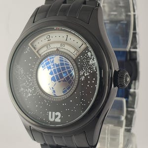 Puede incluir: Reloj de pulsera negro con una esfera &uacute;nica que presenta un dise&ntilde;o de globo terr&aacute;queo y el texto "U2". El reloj tiene una correa negra y una caja redonda negra. La esfera del reloj tiene un dise&ntilde;o de globo terr&aacute;queo blanco y azul.
