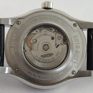 May include: Close-up of the back of a Roamer Roto Date watch. The watch has a silver-colored case with a black leather strap. The watch face is visible, displaying the inner workings, with the text "25 Jewels Swiss Made".