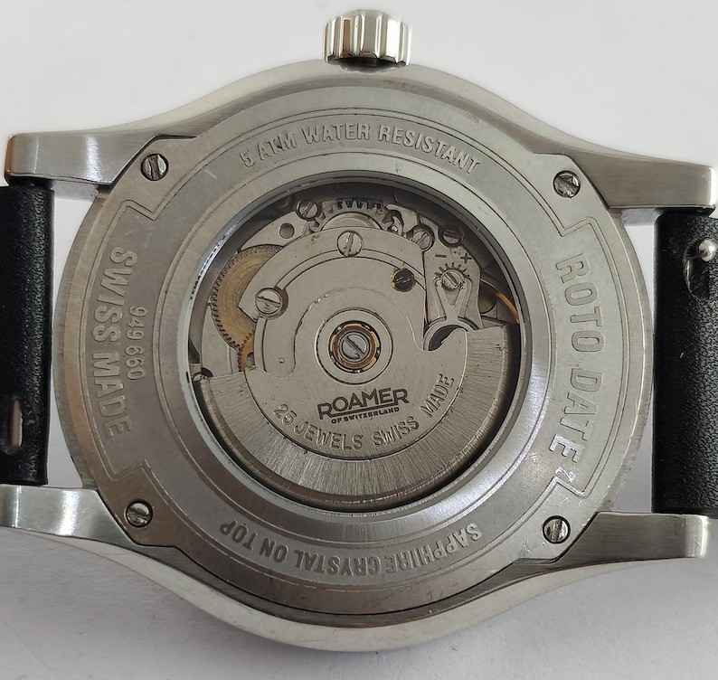May include: Close-up of the back of a Roamer Roto Date watch. The watch has a silver-colored case with a black leather strap. The watch face is visible, displaying the inner workings, with the text "25 Jewels Swiss Made".