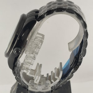 Puede incluir: Un reloj de pulsera negro con una esfera redonda y una correa negra a juego. El reloj se exhibe sobre un soporte de pl&aacute;stico transparente, mostrando su perfil lateral y su intrincado dise&ntilde;o. La correa es texturizada.