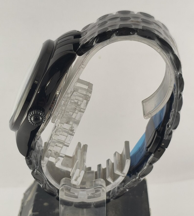 Puede incluir: Un reloj de pulsera negro con una esfera redonda y una correa negra a juego. El reloj se exhibe sobre un soporte de pl&aacute;stico transparente, mostrando su perfil lateral y su intrincado dise&ntilde;o. La correa es texturizada.