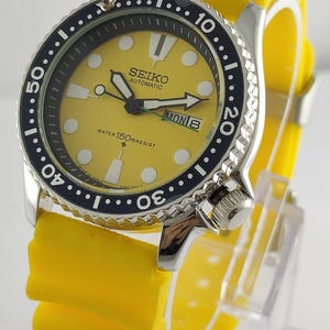 Peut inclure: Une montre Seiko Automatic avec un cadran jaune et une lunette noire rotative. Elle est dotée d'un bracelet en caoutchouc jaune et d'un affichage de la date indiquant "MON 8". La montre est étanche à 150m.