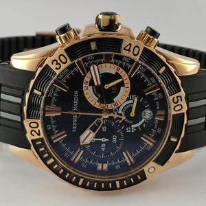 May include: A Ulysse Nardin chronograph watch with a rose gold case, a black dial featuring luminous markers, and a black rubber strap with rose gold inserts. The bezel is black with rose gold numerals, and the watch includes three sub-dials.