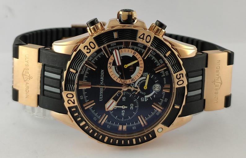 May include: A Ulysse Nardin chronograph watch with a rose gold case, a black dial featuring luminous markers, and a black rubber strap with rose gold inserts. The bezel is black with rose gold numerals, and the watch includes three sub-dials.