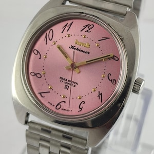 Puede incluir: Reloj de pulsera vintage plateado con esfera rosa. El reloj presenta agujas e indicadores dorados, con el texto "Kohinoor" y "PARA SHOCK 17 JEWELS" visibles. La correa es de metal plateado. La esfera mide aproximadamente 3 cm de diámetro.