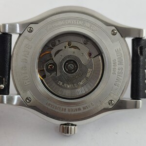 May include: Close-up of the back of a silver-toned watch with a black leather strap. The watch mechanism is visible through a clear glass back. Text includes "SAPPHIRE CRYSTAL ON TOP", "ROTO DATE", "25 JEWELS SWISS MADE", and "5 ATM WATER RESISTANT".