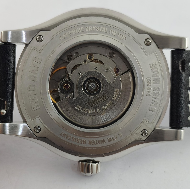 May include: Close-up of the back of a silver-toned watch with a black leather strap. The watch mechanism is visible through a clear glass back. Text includes "SAPPHIRE CRYSTAL ON TOP", "ROTO DATE", "25 JEWELS SWISS MADE", and "5 ATM WATER RESISTANT".