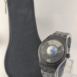 Puede incluir: Reloj de pulsera negro con un dise&ntilde;o &uacute;nico que presenta un globo terr&aacute;queo y un patr&oacute;n de estrellas en la esfera. El reloj tiene una correa de metal negro y el texto "U2" es visible. Tambi&eacute;n hay un estuche de cuero negro con cremallera.
