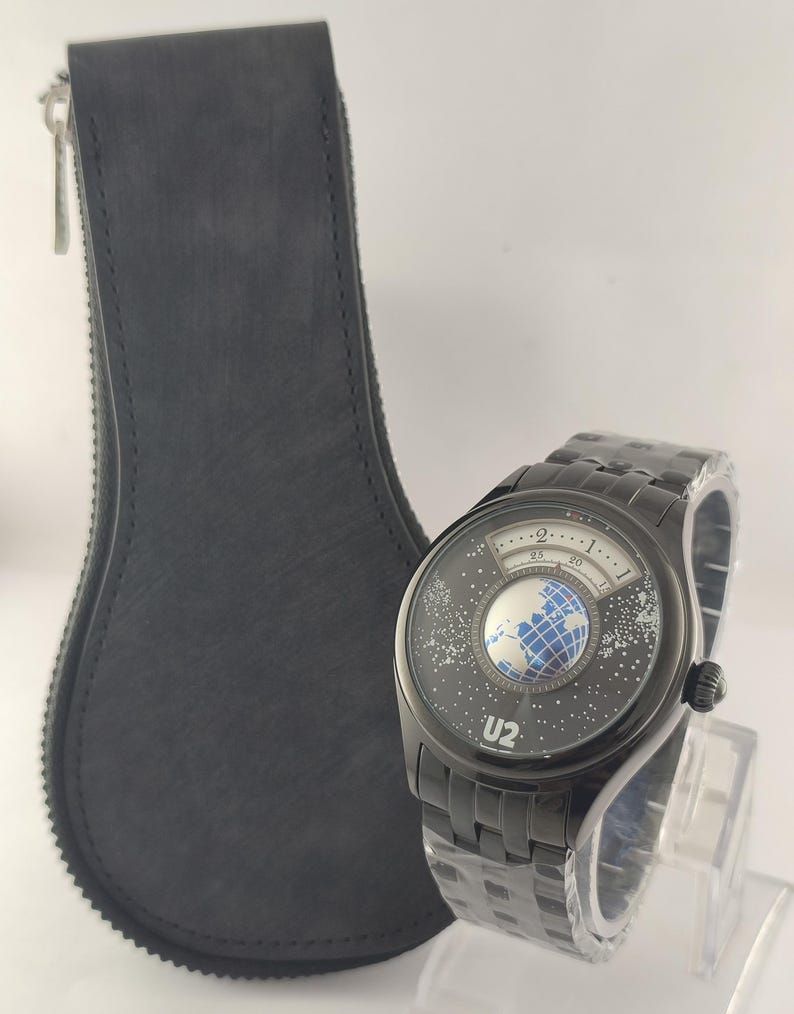Puede incluir: Reloj de pulsera negro con un dise&ntilde;o &uacute;nico que presenta un globo terr&aacute;queo y un patr&oacute;n de estrellas en la esfera. El reloj tiene una correa de metal negro y el texto "U2" es visible. Tambi&eacute;n hay un estuche de cuero negro con cremallera.