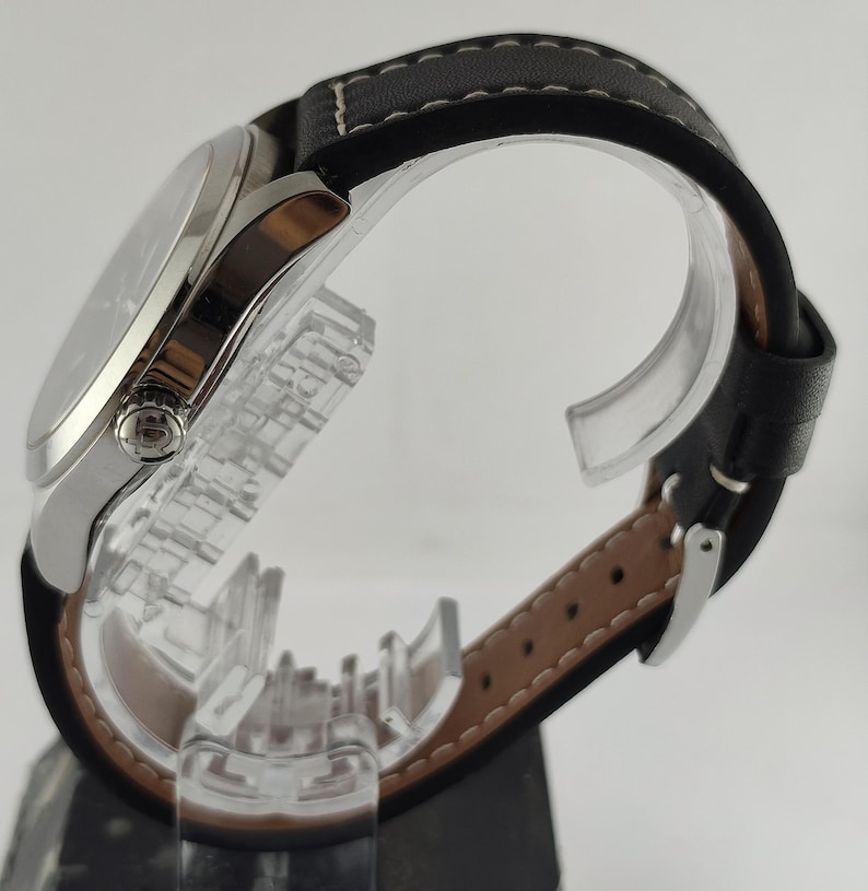May include: A silver-toned wristwatch with a black leather band. The watch face is visible, and the band has white stitching. The watch is displayed on a clear stand, showcasing the side profile and details of the watch.