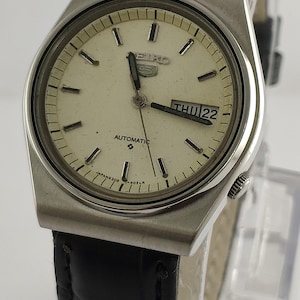 May include: A vintage Seiko 5 automatic wristwatch with a silver case and a cream dial. The watch has a black leather strap and shows the day and date. The watch face has silver markers and hands.