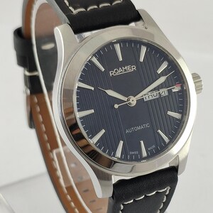 May include: A silver-toned Roamer automatic wristwatch with a black leather band. The watch face is navy blue with silver hands and markers. The band has white stitching. The watch displays the day and date.