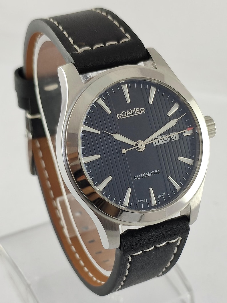 May include: A silver-toned Roamer automatic wristwatch with a black leather band. The watch face is navy blue with silver hands and markers. The band has white stitching. The watch displays the day and date.
