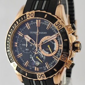 May include: A Ulysse Nardin Marine Diver chronograph watch with a rose gold case, a black rotating bezel with gold numerals, and a dark blue dial. The watch features a black rubber strap with rose gold accents and a date window at the six o'clock position.