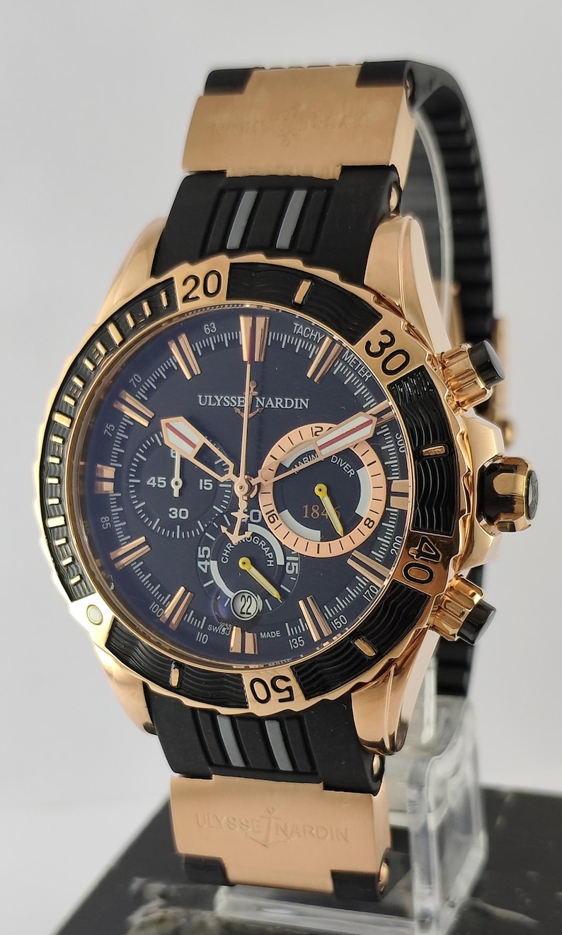 May include: A Ulysse Nardin Marine Diver chronograph watch with a rose gold case, a black rotating bezel with gold numerals, and a dark blue dial. The watch features a black rubber strap with rose gold accents and a date window at the six o'clock position.
