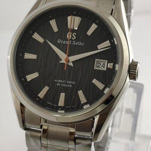 Könnte beinhalten: Eine Grand Seiko Armbanduhr aus Edelstahl mit einem schwarzen, strukturierten Zifferblatt, silbernen Zeigern und einer Datumsanzeige bei 3 Uhr. Die Uhr trägt die Aufschrift "Grand Seiko" und "Hi-Beat 36000 80 Hours" auf dem Zifferblatt.