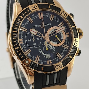 May include: A Ulysse Nardin chronograph watch with a rose gold case and a black rubber strap. The dark blue dial features rose gold hour markers, three sub-dials, a date window at the six o'clock position, and a rotating bezel with gold numerals.