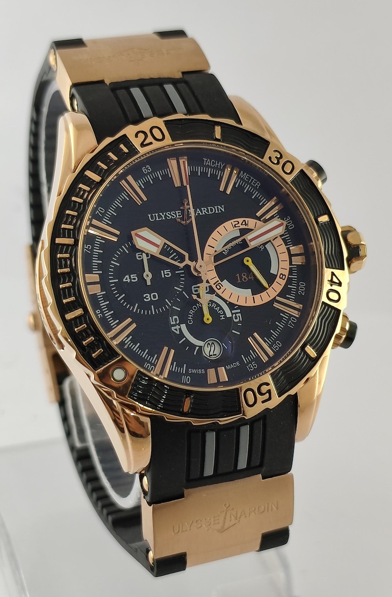 May include: A Ulysse Nardin chronograph watch with a rose gold case and a black rubber strap. The dark blue dial features rose gold hour markers, three sub-dials, a date window at the six o'clock position, and a rotating bezel with gold numerals.