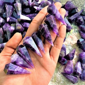May include: A collection of faceted amethyst crystal points in varying shades of purple and violet. The crystals are held in a hand, with many more scattered in the background. The crystals have a geometric shape with a pointed tip.