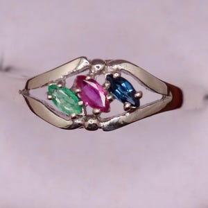 May include: A silver-toned ring featuring a bypass-style band with three marquise-cut gemstones set in the center. The stones are arranged horizontally, displaying a vibrant green, a deep pink, and a dark blue color in a polished metal setting.
