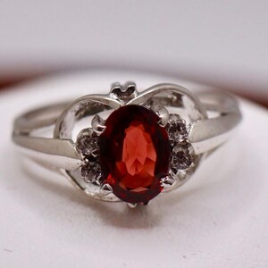 May include: A silver-toned ring featuring a central oval-cut deep red gemstone. The setting includes small clear accent stones on either side of the center gem, integrated into a polished metal band with a split-shank design at the shoulders.