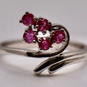May include: A silver-toned ring featuring a bypass-style band that curves upward to hold a cluster of five small, round, vibrant pinkish-red gemstones. The metal band has a polished, reflective finish and tapers as it wraps around the finger.