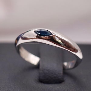May include: A silver ring with a dark blue, oval-shaped stone. The ring is displayed on a black stand, with a blurred background featuring a blue and white floral design.