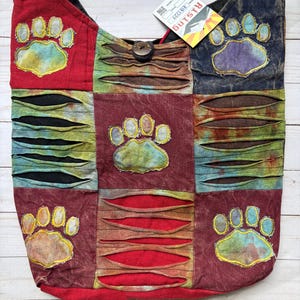 May include: A patchwork hobo bag featuring a red, dark blue, and maroon color scheme with horizontal slashed fabric details. The bag is decorated with five embroidered paw print patches in yellow, blue, and green. A wooden button closure is at the top.