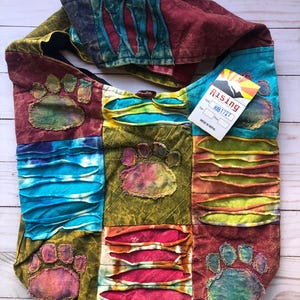 May include: A colorful patchwork tote bag with a tie-dye pattern and paw print designs. The bag has a black strap and a tag that reads "Rising" and "RIB 1727". Made in Nepal.